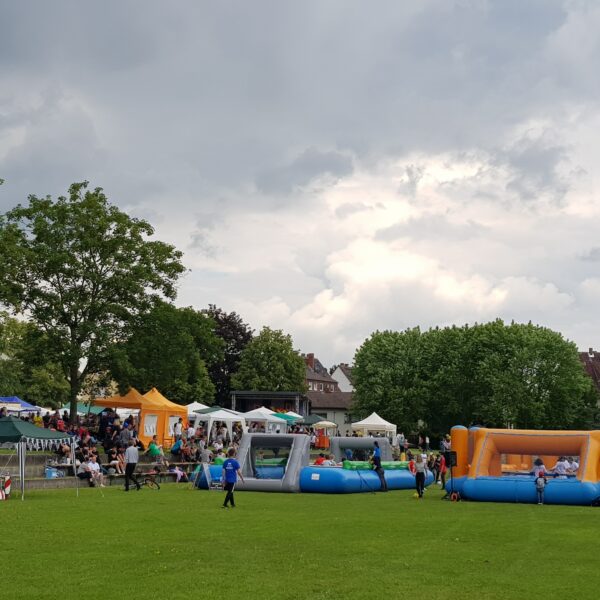 Seewiese Friedberg. Menschenkickercup und Fest der Kulturen. Eine Bühne, viele Pavillons und zwei Kicker Felder sind aufgebaut. Menschen sind auf dem Platz. Bäume säumen die grüne Wiese.