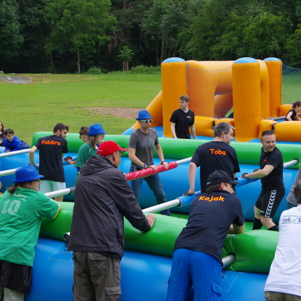 Mehrere Personen stehen auf dem Menschenkicker-Feld und spielen Fußball. Zuschauer säumen das Spielfeld.