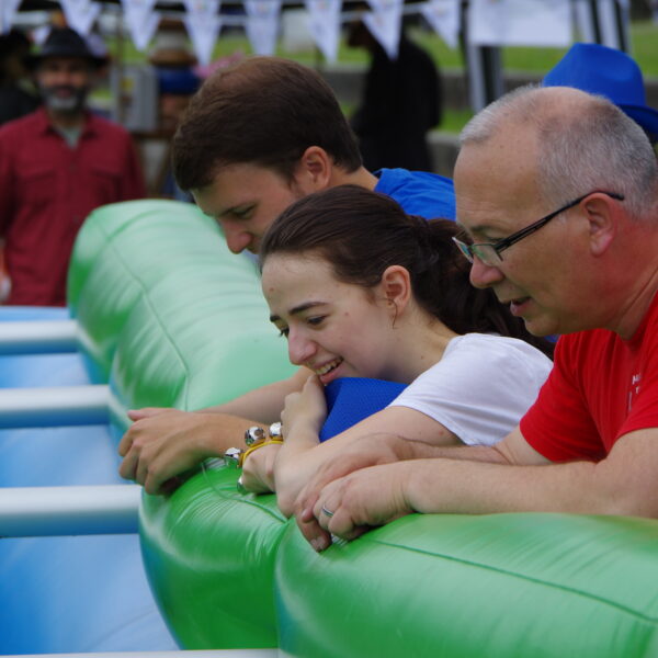 Menschenkicker Cup. Drei Zuschauer stützen sich nach vorne auf dem Menschenkicker ab und verfolgen gespannt das Spiel