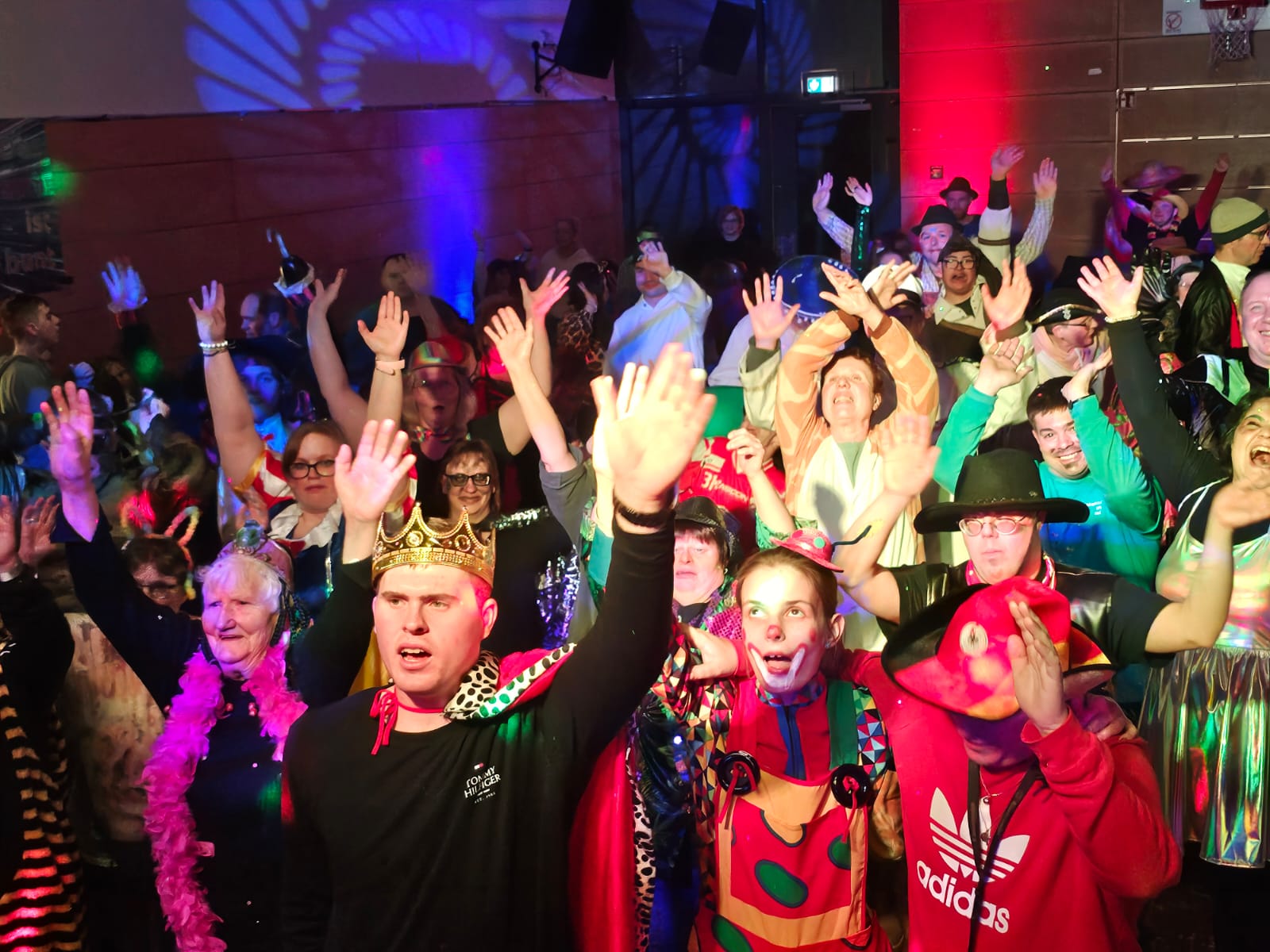 Viele Menschen sind in einem Raum. Sie feiern gemeinsam Fasching. Der Raum ist bunt beleuchtet. Die Menschen feiern. Sie sind verkleidet. Die Menschen sind fröhlich und heben die Hände in die Luft.