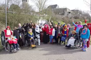 Faschingsumzug. Die Laufgruppe von Mensch mach mit hat sich für ein Foto aufgestellt. Im Hintergrund ist das Wahrzeichen von Friedberg, der Adolfsturm zu sehen. Auf dem Bild sind viele Menschen in bunten Kostümen zu sehen. Auch drei Rollstuhlfahrer sind mit dabei.