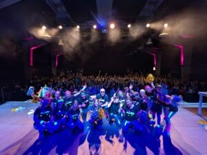 Die Schlagerfamily, insgesamt 17 Personen, macht ein Abschlussfoto auf einer großen Bühne. Im Hintergrund ist das Publikum zu sehen. Fast 800 Leute, die in die Kamera Jubeln.